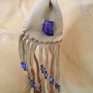 May include: A small, tan leather pouch with a purple stone and blue beads hanging from the bottom. The pouch has a fringe and is tied with a brown leather cord.
