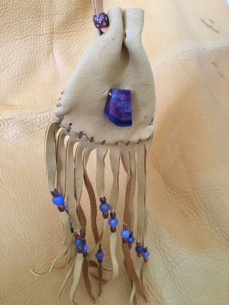 May include: A small, tan leather pouch with a purple stone and blue beads hanging from the bottom. The pouch has a fringe and is tied with a brown leather cord.