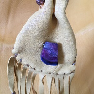 May include: A small, light brown leather pouch with a purple and blue agate stone attached. The pouch has a fringe and blue beads on the bottom.