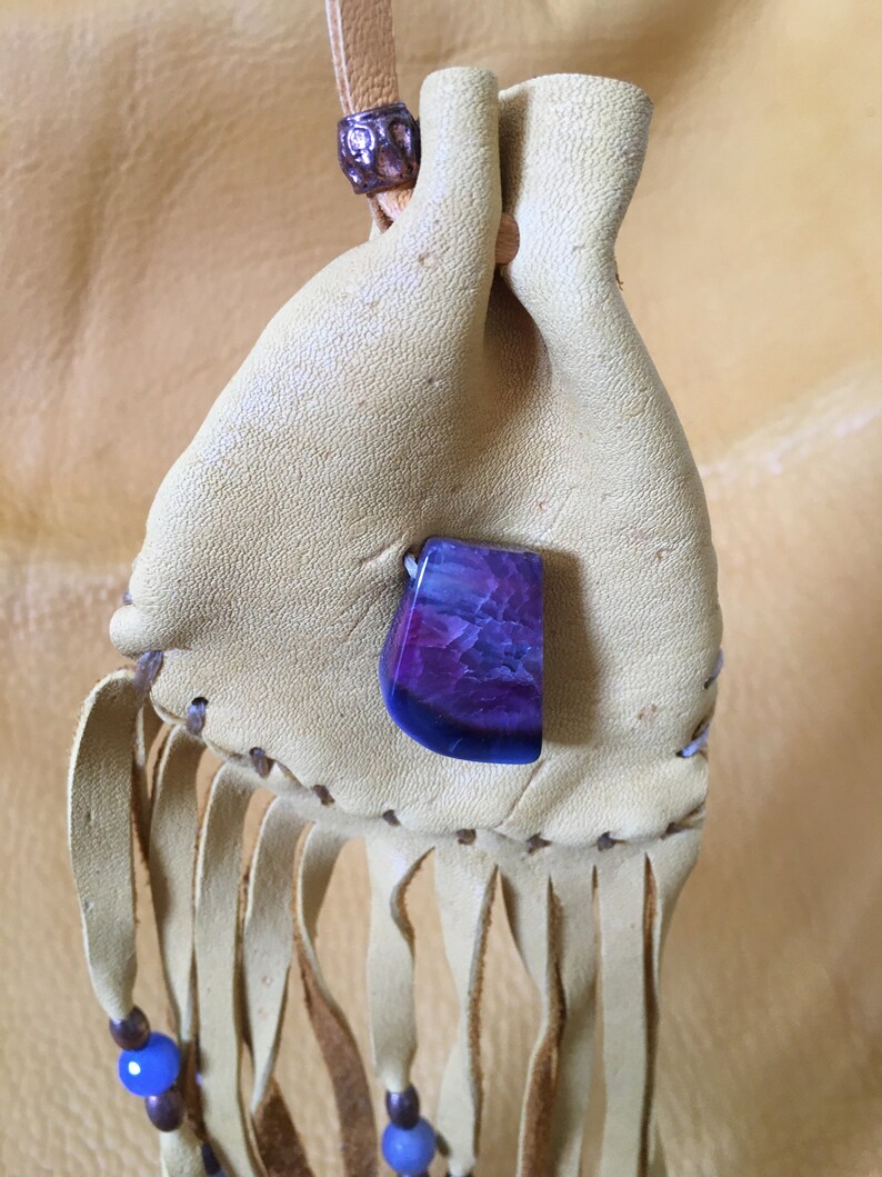 May include: A small, light brown leather pouch with a purple and blue agate stone attached. The pouch has a fringe and blue beads on the bottom.