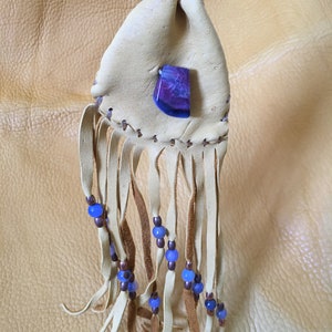 May include: A small, light brown leather pouch with a purple and white agate stone attached. The pouch has a fringe with blue beads and brown leather ties.