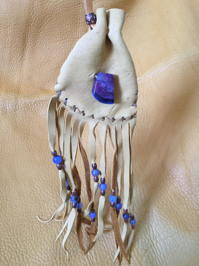 May include: A small, light brown leather pouch with a purple and white agate stone attached. The pouch has a fringe with blue beads and brown leather ties.