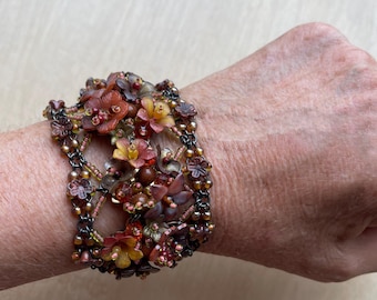 May include: A floral bracelet with a mix of red, orange, and yellow flowers. The bracelet is made of beads and small flowers, creating a textured look. The bracelet is worn on a wrist.