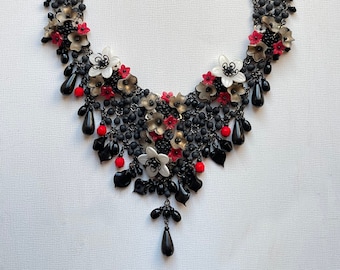 May include: A black beaded necklace with a cascading design featuring red, white and gold flowers. The necklace has a black and silver clasp.