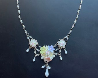 May include: A delicate silver necklace with a floral pendant. The pendant features a cluster of pastel pink, yellow, and blue flowers with a single white teardrop-shaped crystal hanging below.