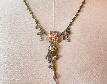 May include: A delicate necklace with a floral pendant made of pink, green and purple beads. The necklace is made of a silver chain with small green beads.