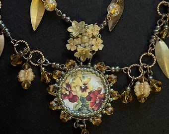 May include: A gold necklace with a floral cameo pendant, featuring a cluster of white flowers and a round cameo with a floral design. The necklace is adorned with gold leaves and amber-coloured beads.