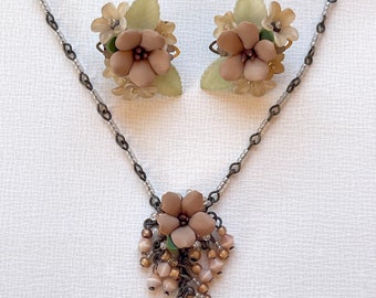 Op de afbeelding: Een delicate set met een ketting en oorbellen met een bloemenmotief met bruine en groene accenten. De ketting heeft een hanger met een cluster van kleine kralen en een grotere bloemenhanger. De oorbellen zijn van het clip-on type met een enkele bloemenhanger.