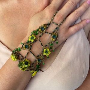 May include: A delicate gold chain bracelet with green and yellow flower accents. The bracelet is designed to fit around the wrist and fingers, with a chain that extends up the arm.