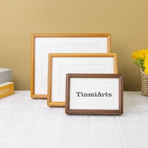 Puede incluir: Tres marcos de fotos de madera de diferentes tamaños. El marco más pequeño es marrón oscuro y tiene el texto "TinmiArts" impreso en el paspartú blanco. Los dos marcos más grandes son de color marrón claro.