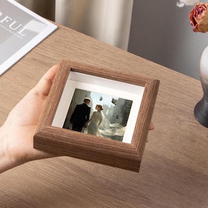Puede incluir: Un marco de fotos cuadrado de madera con una foto de boda. El marco es de color marrón medio y tiene un diseño simple y clásico. La foto muestra a una novia y un novio. El marco se sostiene en una mano.