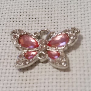 Peut inclure: Broche argentée en forme de papillon ornée de pierres roses. La broche présente un design symétrique avec des pierres claires en forme de cristal soulignant les ailes et le corps, et des pierres roses au centre.