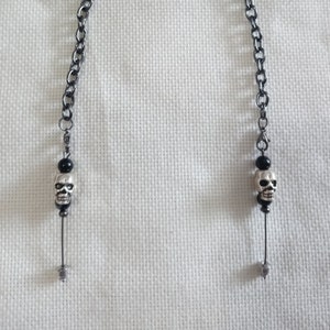 May include: A silver-toned chain with a skull charm at each end. The chain is made of small, oval links. Each charm features a small skull with black bead accents. The chain is displayed against a white background.