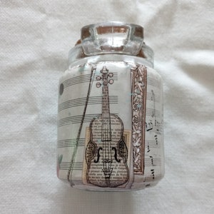 May include: A small, clear glass jar with a decorative paper wrap featuring a violin illustration, musical notes, and text. The jar has a clear glass stopper and a cork. The overall design has a vintage, musical theme.