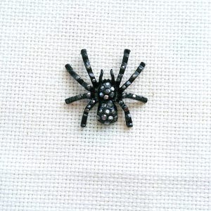 Peut inclure: Une broche en forme d'araignée noire avec une finition métallique, ornée de nombreux petits cristaux sombres et réfléchissants sur le corps et les huit pattes. L'araignée est représentée dans une position symétrique et plate.