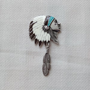 May include: A silver-toned metal pin in the shape of a Native American chief's head, with a feather pendant. The pin features a headdress with white, brown, and turquoise accents. The feather hangs below the head.