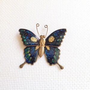 Puede incluir: Un broche de mariposa con cuerpo y antenas de metal dorado. Las alas están rellenas con un acabado de esmalte moteado en azul oscuro, verde y negro, acentuado por dos puntos de purpurina dorada en las secciones superiores de las alas.