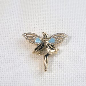 May include: A gold-coloured fairy brooch with rhinestone-studded wings and light blue accents. The fairy is depicted in a flowing dress with a delicate design. The brooch is set against a white background.