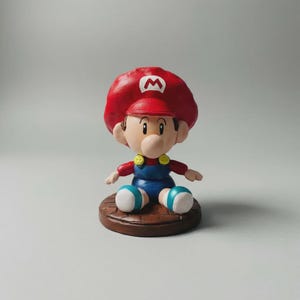 Puede incluir: Una figura de Baby Mario, personaje de la serie de videojuegos Super Mario. La figura lleva un gorro rojo con un emblema blanco, una camisa roja, un peto azul y zapatos blancos y azules. Está sentado sobre una base marrón.