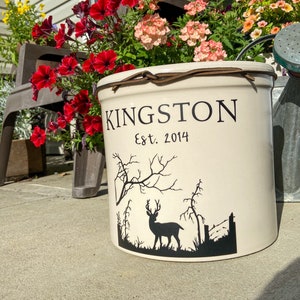 May include: A white ceramic planter with a brown leather strap around the top. The planter has a black silhouette of a deer in a forest scene and the text "KINGSTON Est. 2014".