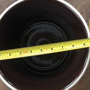 May include: A brown ceramic crock with a white rim, measuring 9 inches in diameter. The crock is sitting on a wooden surface.