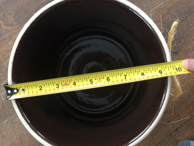 May include: A brown ceramic crock with a white rim, measuring 9 inches in diameter. The crock is sitting on a wooden surface.