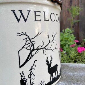 May include: White ceramic crock with a black leather strap and a black silhouette of a deer and trees. The crock has the word "WELCOME" written on the front.
