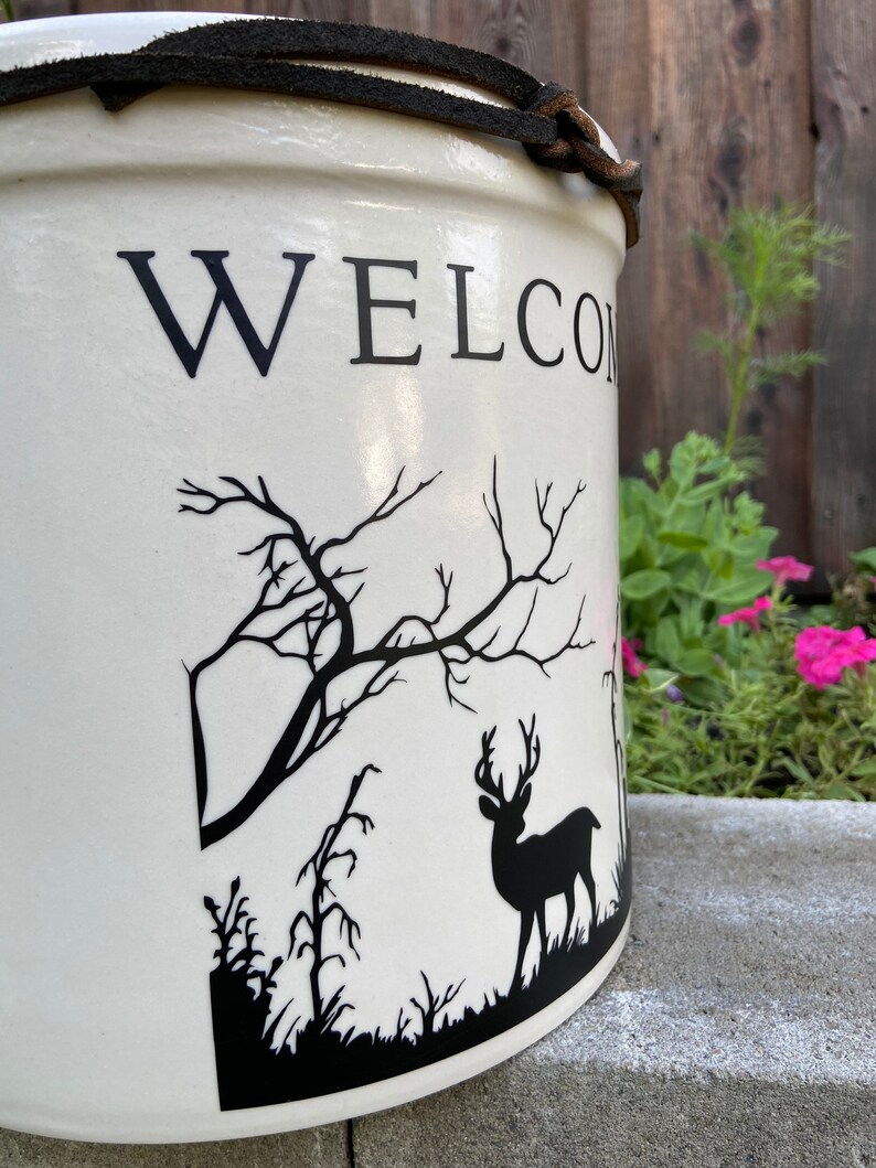 May include: White ceramic crock with a black leather strap and a black silhouette of a deer and trees. The crock has the word "WELCOME" written on the front.