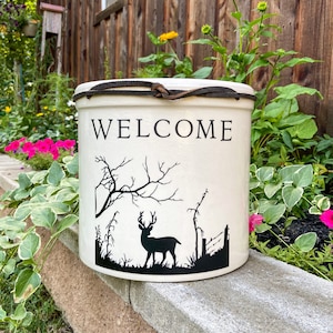 May include: White ceramic crock with a brown leather strap and a black silhouette of a deer in a woodland scene. The crock has the word "WELCOME" written on the front.