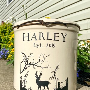 May include: White ceramic crock with a brown leather strap around the top. The crock has a black silhouette of a deer in a forest scene and the text "HARLEY Est. 2015".