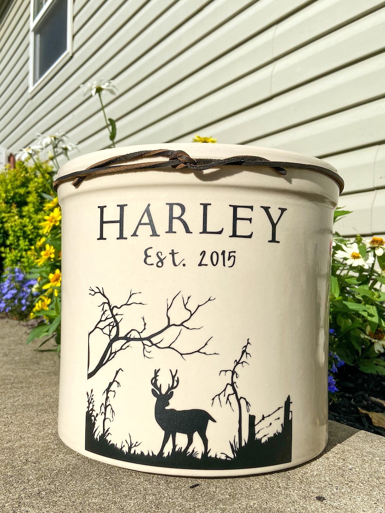 May include: White ceramic crock with a brown leather strap around the top. The crock has a black silhouette of a deer in a forest scene and the text "HARLEY Est. 2015".