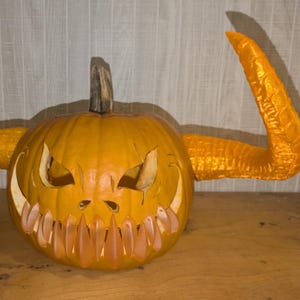 Peut inclure: Une citrouille orange sculptée avec un visage menaçant et de grandes cornes dorées incurvées. La citrouille a des yeux triangulaires et un sourire denté et irrégulier, parfait pour Halloween.