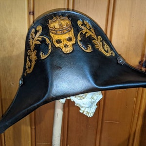 May include: Black leather tricorn hat with gold skull and crown design. The hat features ornate gold scrollwork and is mounted on a skull. A unique, decorative accessory.