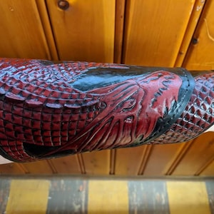 May include: A red and black leather arm guard with a dragon design. The guard is made of embossed leather and has a textured surface.