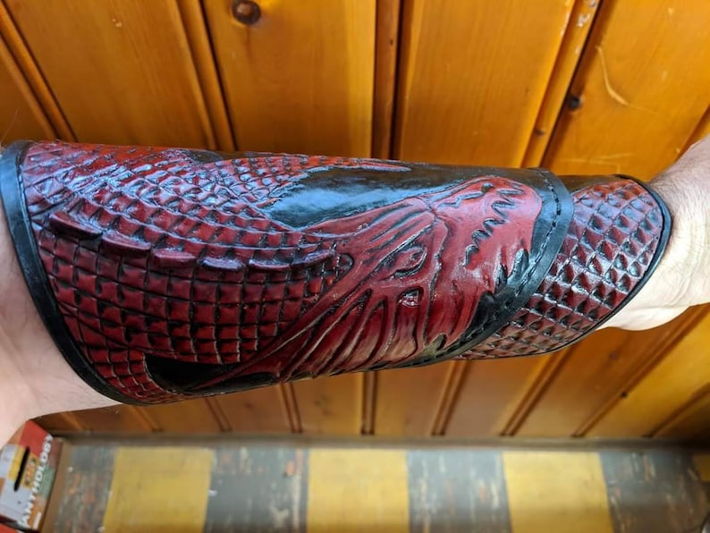 May include: A red and black leather arm guard with a dragon design. The guard is made of embossed leather and has a textured surface.