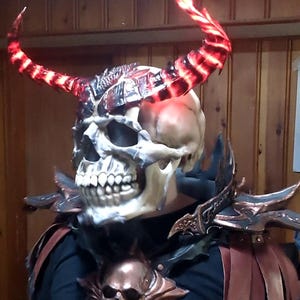 Op de afbeelding: Een schedelmasker met gloeiende rode hoorns en een gedetailleerd metalen borststuk met een schedelontwerp.