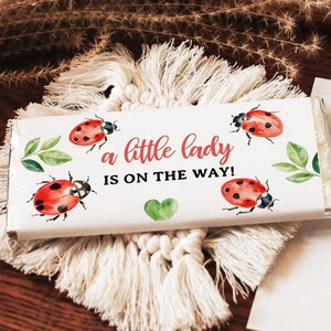 May include: White chocolate bar with a red and black ladybug design. The text on the bar reads "a little lady is on the way!"