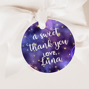 Puede incluir: Una caja de regalo blanca atada con una cinta blanca, con una etiqueta redonda. La etiqueta tiene un diseño de galaxia azul y púrpura, con el texto "a sweet thank you LOVE, Luna" en escritura blanca.
