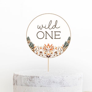 Op de afbeelding: Een witte cake met een ronde caketopper waarop in bruin schrift "wild one" staat. De caketopper is versierd met bruine, oranje en groene bloemmotieven.