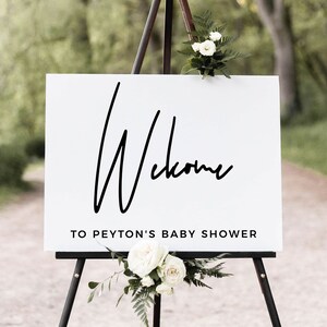 May include: A white sign with black text on an easel in a forest setting. The sign reads "Welcome to Peyton's Baby Shower".
