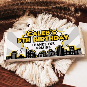 May include: A white chocolate bar wrapper with a yellow and black comic book style design. The wrapper says "Caleb's 5th Birthday" and "Thanks for coming".