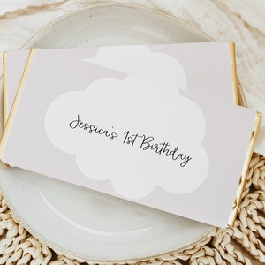 Op de afbeelding: Een lichtroze kaart met gouden randen en een witte wolkvorm met de tekst "Jessica's 1st Birthday" in zwarte cursieve letters.