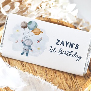 Puede incluir: Un envoltorio de chocolate blanco con una ilustración de un astronauta azul y blanco que sostiene globos con forma de planetas. El texto "ZAYN'S 1st Birthday" está impreso en el envoltorio.