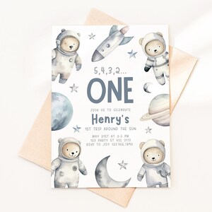 May include: A white invitation with a blue and gray space theme. It features a rocket, planets, stars, and four bears dressed as astronauts. The text reads "5, 4, 3, 2... ONE Join us to celebrate Henry's 1st Trip Around the Sun May 21st at 2-5 PM 123 Party St VIC 3112 RSVP to Joy 1234643890"