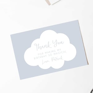 Może przedstawiać: Jasnoniebieska kartka z podziękowaniami z chmurką. Na karcie widnieje napis "Thank You for making my birthday so magical, Love, Patrick." Białe kwiaty i artykuły papiernicze ułożone wokół kartki na białej powierzchni.
