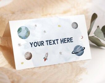 以下が含まれることがあります： 水彩画の宇宙をテーマにした折り畳みカード。カードには惑星、星、ロケットのイラストが描かれています。「YOUR TEXT HERE」の文字は濃いネイビーで印刷されています。カードは白い表面に置かれ、金のフォークが添えられています。