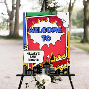 May include: A welcome sign for a baby shower with a comic book theme. The sign is red, yellow, blue, and green with a cityscape and the words "Welcome to Hillary's Baby Shower" and "Wow!" in a speech bubble.