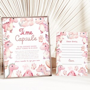 May include: A pink and white time capsule with a dinosaur theme. The text on the card reads "Time Capsule To Be Opened When Emily Turns 18 in 2039".