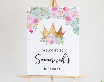 May include: A white sign on a wooden easel with the text "WELCOME TO Savannah's BIRTHDAY!". The sign features a gold crown and floral design in pink, blue, and gold. Perfect for a birthday party.