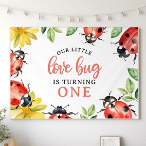 May include: A framed print featuring watercolor illustrations of ladybugs, yellow flowers, and green leaves. The text "Our Little Love Bug Is Turning One" is written in a playful font. The artwork is displayed on a white wall above a wooden table.
